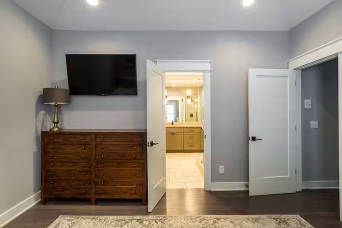Bright modern bedroom with gray walls, dark hardwood floors and area rug, wooden dresser with lamp and wall-mounted TV, and an open door leading to a light-tiled en-suite bathroom with vanity.