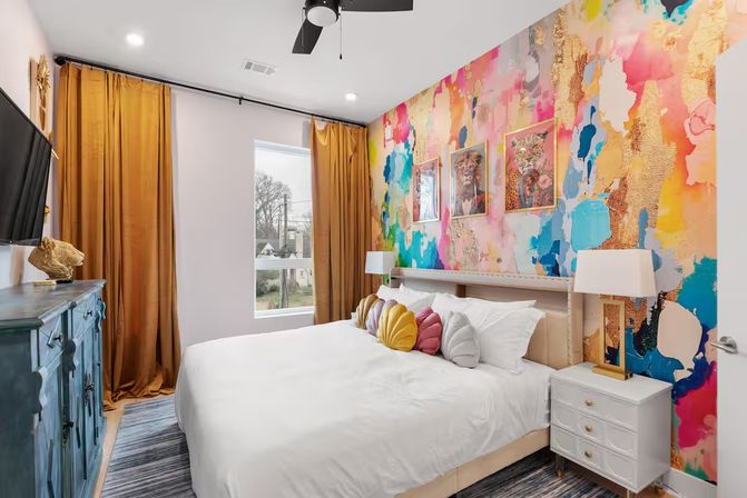 Vibrant modern bedroom with a colorful abstract mural accent wall, white bed topped with shell-shaped throw pillows, gold floor-to-ceiling curtains, teal dresser, white nightstand with lamp, ceiling fan, and window overlooking a neighborhood.