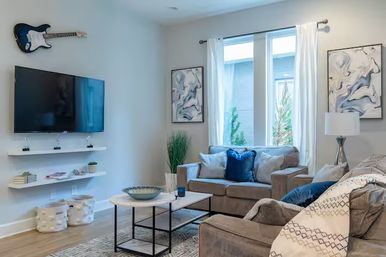 Sunlit modern living room with gray sofas and blue accent pillows, wall-mounted TV and decorative guitar, abstract art, white curtains, coffee table, potted plant and hardwood floor