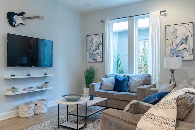 Sunlit modern living room with gray sofas and blue accent pillows, wall-mounted TV and decorative guitar, abstract art, white curtains, coffee table, potted plant and hardwood floor
