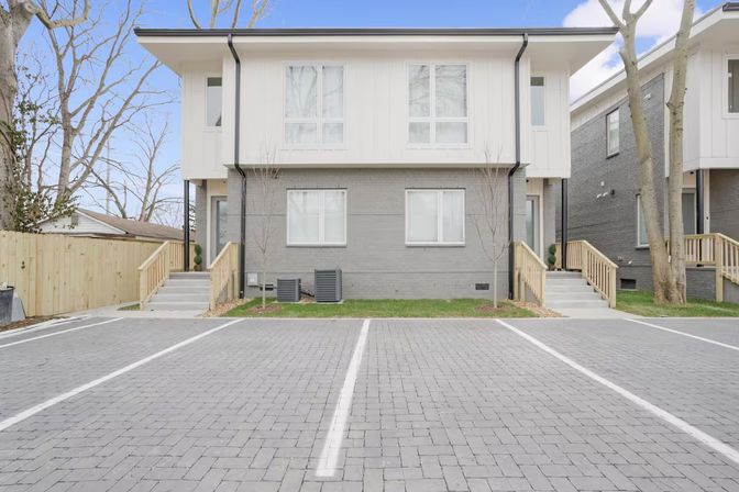 Modern two-story suburban duplex townhouse with gray brick lower level, white siding upper, matching wooden stair entrances and paved parking spaces in front.