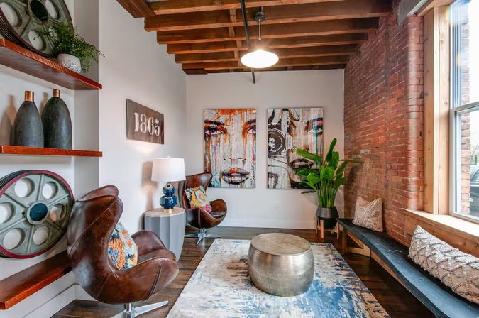 Cozy industrial loft lounge with exposed brick and wood-beam ceiling, two brown leather swivel chairs with colorful pillows, abstract face diptych, round hammered metal coffee table on blue rug, window bench, potted plant and decorative shelves
