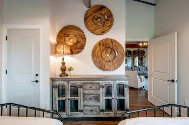 Cozy modern farmhouse bedroom interior: distressed gray sideboard with lamp and small plant, three large reclaimed wooden round wall discs above, metal bed frames in foreground and open doorway to a living room.