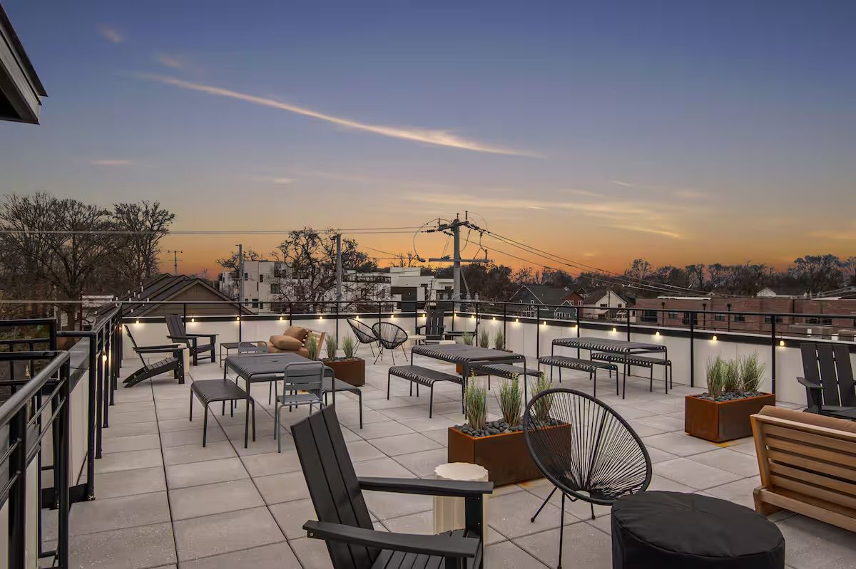 Modern rooftop terrace at sunset with cozy outdoor lounge seating, metal and wooden chairs, benches, planters and string lights along a glass railing overlooking a residential neighborhood skyline
