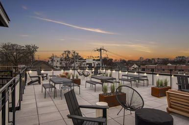 Modern rooftop terrace at sunset with cozy outdoor lounge seating, metal and wooden chairs, benches, planters and string lights along a glass railing overlooking a residential neighborhood skyline