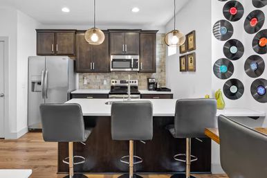 Modern open-concept kitchen with white island and three gray bar stools, dark wood cabinets, stainless steel fridge and oven, pendant lights, stone tile backsplash and vinyl record accent wall for a retro vibe.