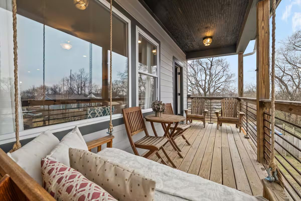 Cozy covered wooden porch deck with a rope-hung daybed and patterned pillows, small round bistro table with folding chairs, and two wooden lounge chairs overlooking a suburban backyard with bare trees.
