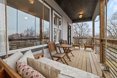 Cozy covered wooden porch deck with a rope-hung daybed and patterned pillows, small round bistro table with folding chairs, and two wooden lounge chairs overlooking a suburban backyard with bare trees.