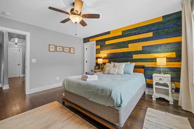 Cozy modern bedroom interior with teal bedding, bold yellow-and-green horizontal wood plank accent wall, hardwood floors, ceiling fan and bedside tables with lamps.