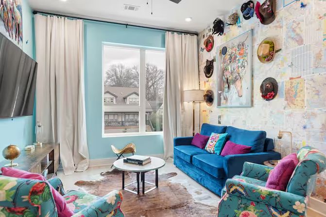 Vibrant boho living room with teal walls, blue velvet sofa, floral armchairs, map-print wallpaper with mounted hats, cowhide rug, round coffee table and large window overlooking suburban houses.