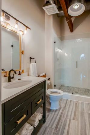 Modern farmhouse-style bathroom with dark vanity and brass pulls, white countertop and black faucet, glass-enclosed walk-in shower, exposed wood beam ceiling and gray wood-look tile floor.