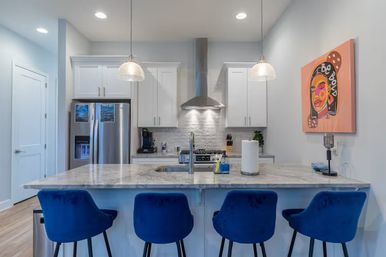 Bright modern kitchen with marble island and breakfast bar lined with four royal blue velvet stools, white cabinets, stainless steel fridge and range hood, two glass pendant lights and colorful pop-art portrait on the wall.