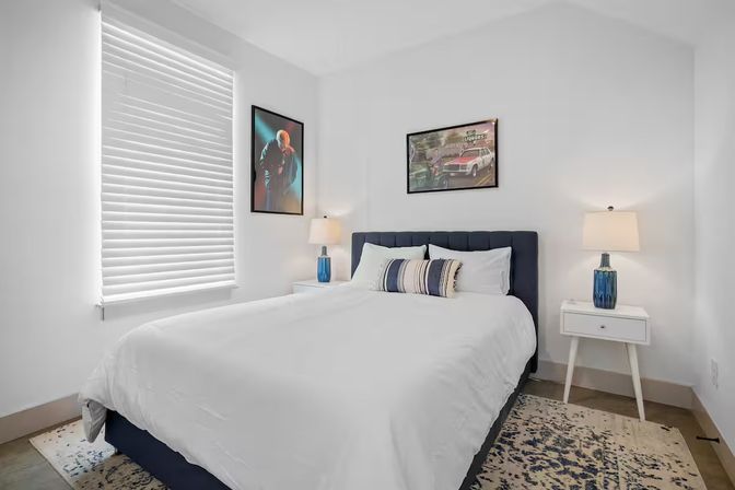 Bright modern bedroom interior with a navy upholstered headboard, crisp white bedding and striped accent pillows, two white nightstands with blue glass lamps, framed wall art, window blinds and a patterned area rug.