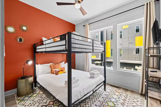 Cheerful modern apartment bedroom with black metal bunk bed and white linens, bold orange accent wall with sunburst mirrors, patterned rug, floor lamp, and large windows overlooking a building with yellow panels.