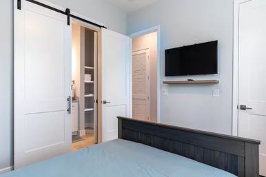 Modern bedroom with dark wood bed and blue bedding, white sliding barn door opening to a linen closet and ensuite bathroom, wall-mounted flat-screen TV on soft gray walls with a floating shelf