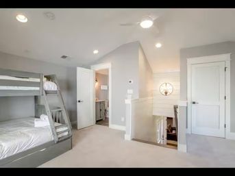 Bright upstairs landing bedroom featuring gray wooden bunk beds with white linens and folded towels, open en-suite bathroom door, carpeted floor, stairwell railing, ceiling fan and recessed lighting.