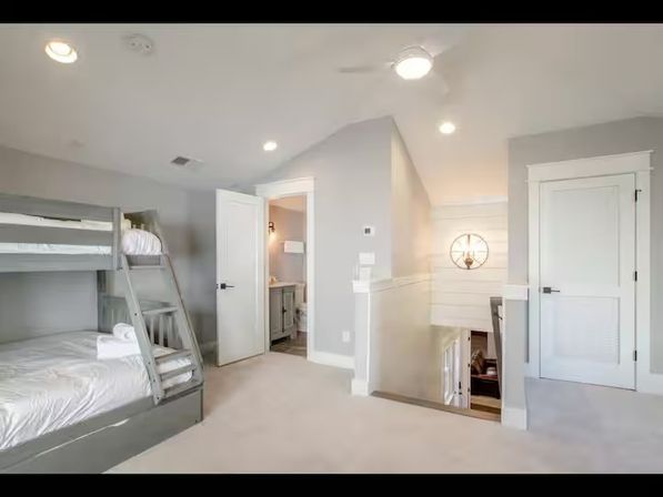 Bright upstairs landing bedroom featuring gray wooden bunk beds with white linens and folded towels, open en-suite bathroom door, carpeted floor, stairwell railing, ceiling fan and recessed lighting.