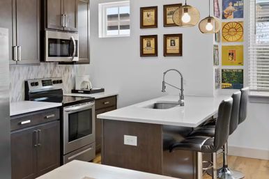 Modern urban kitchen with white quartz island and stainless sink, dark wood cabinets, stainless steel stove and microwave, leather bar stools, woven pendant lights and framed vintage posters by a sunny window.