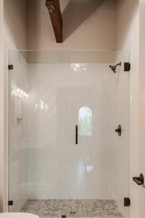 Modern farmhouse frameless glass shower enclosure with white hexagon wall tile, mosaic floor tile, oil‑rubbed bronze fixtures and exposed wooden ceiling beam