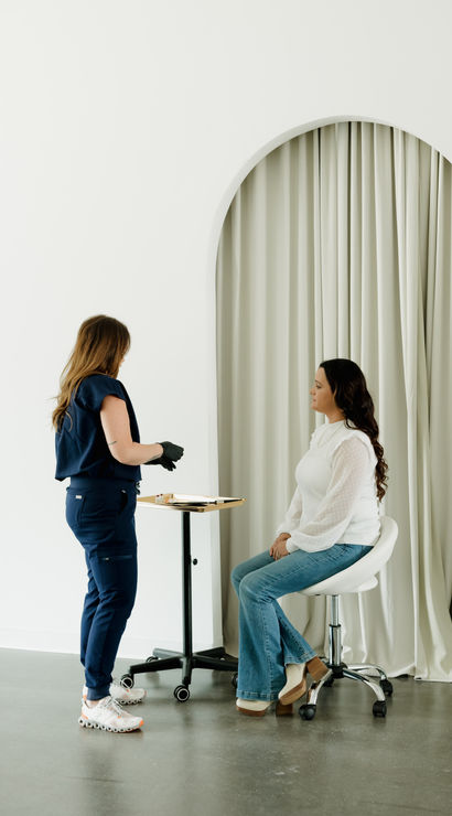Minimalist clinic consultation with a seated female patient on a stool and a gloved practitioner standing beside a tray, set against a white arched curtain backdrop.