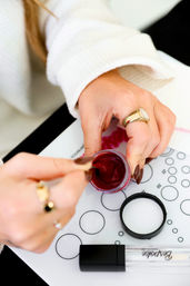 Close-up of hands stirring deep burgundy gel polish with a brush on a circular mixing mat, manicure tools, gold rings and white sweater sleeve visible — nail salon nail art setup.
