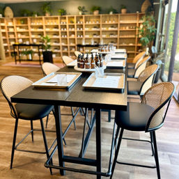 Bright boutique scent workshop: long black table set with trays, glass cups and amber dropper bottles, rattan-back chairs and wooden shelving filled with jars in a light-filled studio.