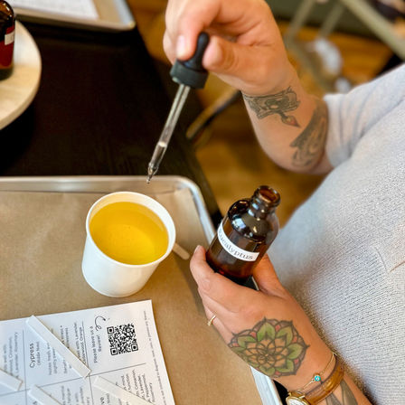 Tattooed hands dripping eucalyptus oil from a dropper into a yellow paper cup during a DIY aromatherapy blending session, tray and QR handout visible.
