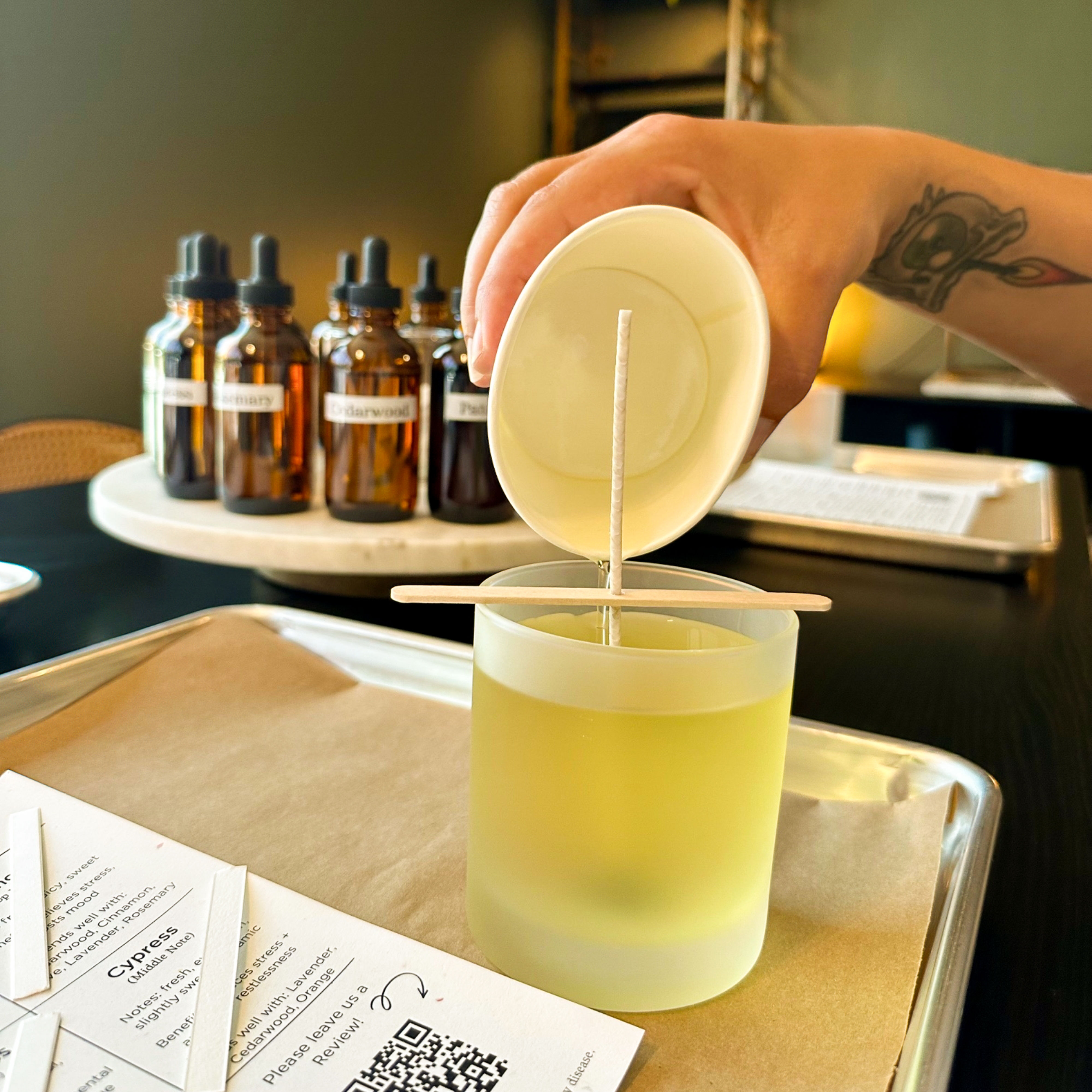 Close-up of a DIY candle-making scene: a tattooed hand pours pale yellow melted wax from a paper cup into a frosted glass jar while a wick is held centered by a wooden stick; amber fragrance dropper bottles and scent sample cards sit on the tabletop.