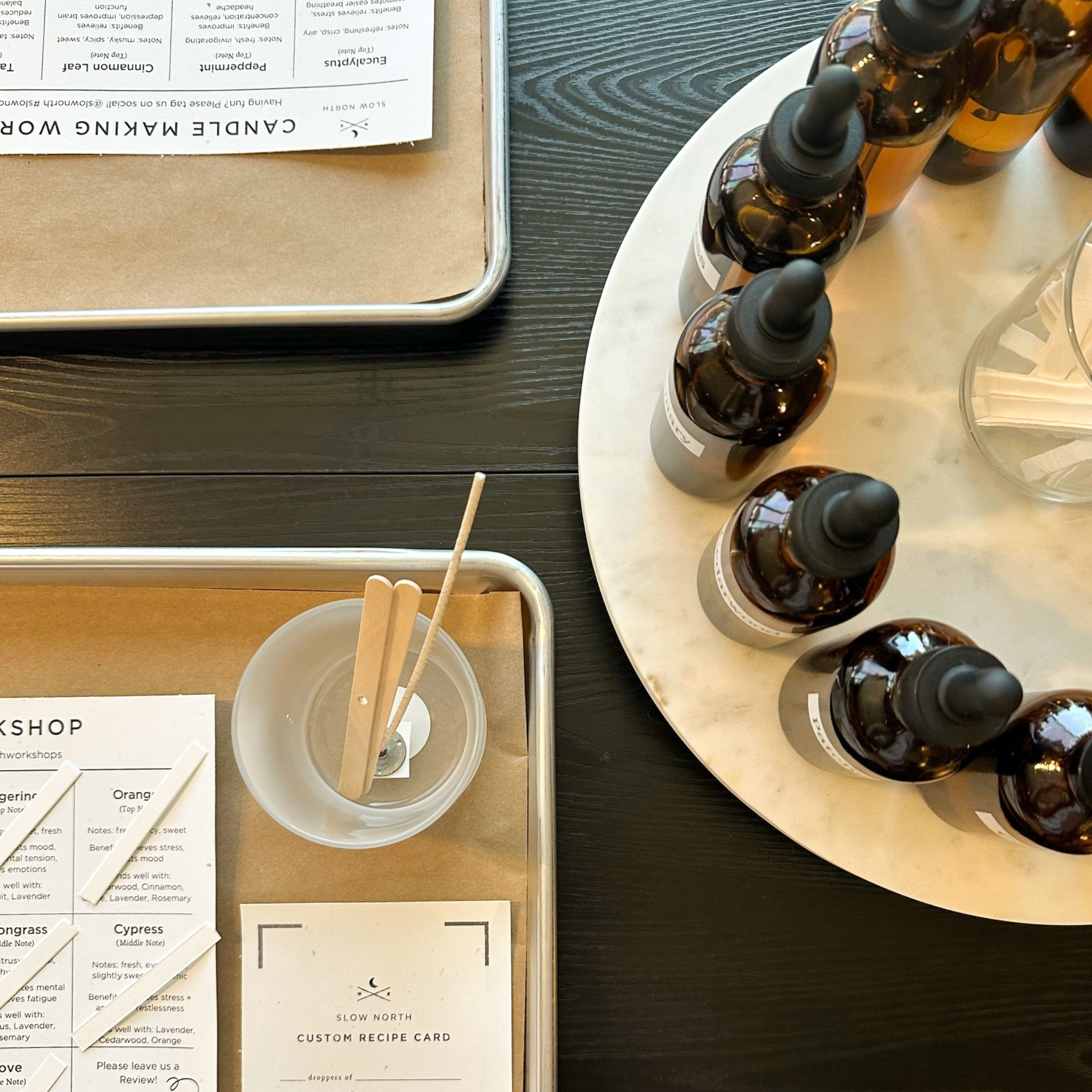 Overhead view of a candle-making workshop setup on a dark wood table: amber glass dropper bottles on a round marble tray, metal trays with custom recipe cards and scent strips, and a plastic cup holding wooden stir sticks — ready to mix scents.