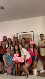 Barbie-themed group photo in a living room: friends in pink outfits smiling, one holding a pink Barbie sign and another wearing a pink cowboy hat.