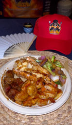 Mouthwatering hibachi surf-and-turf plate with grilled lobster tail, buttered shrimp and scallops, seared steak cubes and stir-fried broccoli and onions on a woven placemat, folding fan and red cap in background.