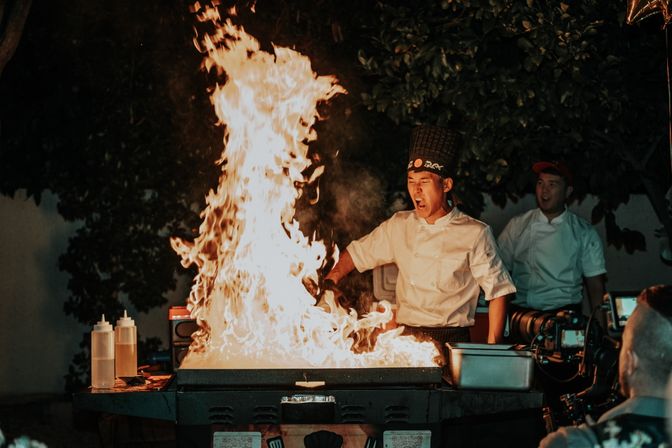 Hibachi-style chef in white jacket and tall hat creating a towering flame on an outdoor teppanyaki grill during a nighttime cooking show, with assistants and diners watching.