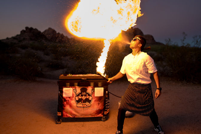 Chef in white shirt and striped apron breathes a giant fireball over a mobile grill cart at dusk, the orange plume lighting up a rocky desert background.