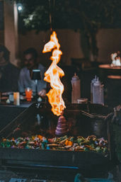 Teppanyaki grill spectacle with a flaming onion “volcano” erupting bright orange flame on a hot griddle, surrounded by colorful chopped vegetables and a chef’s gloved hand with spatula in a dim restaurant with blurred diners.