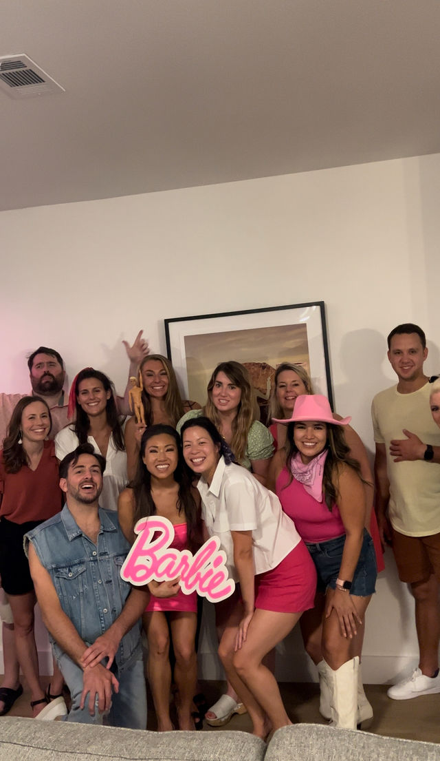Barbie-themed house party in a living room — a large group of smiling friends wearing pink outfits and a pink cowboy hat, holding a 'Barbie' sign and toy doll in front of framed wall art.