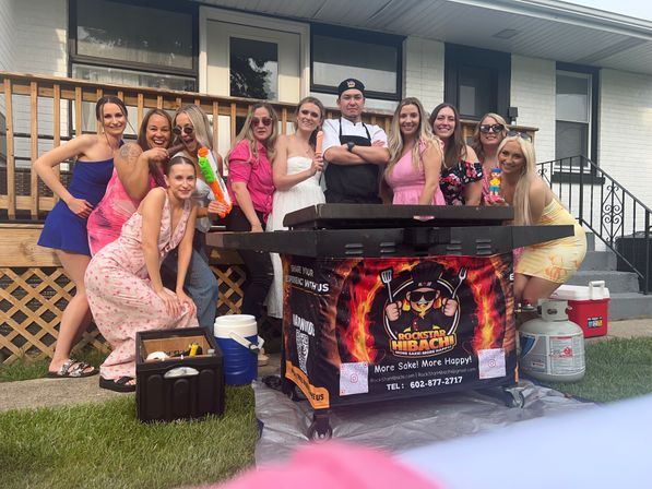 Backyard summer party: group of women in colorful dresses and a chef posing behind a mobile hibachi grill on a suburban front porch, smiling with playful props.