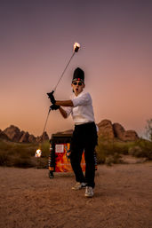 Chef in tall hat and sunglasses spinning flaming chains during an outdoor desert sunset performance with rocky formations in the background