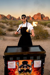 Chef in apron and headband twirling flaming torches over an outdoor griddle at a desert sunset with red rock formations in the background — energetic food pop-up scene.