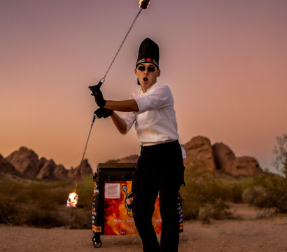Fire-spinning chef performer in white jacket and tall hat twirls a flaming chain in a desert at sunset, wearing gloves and sunglasses with rocky hills and a food cart in the background.