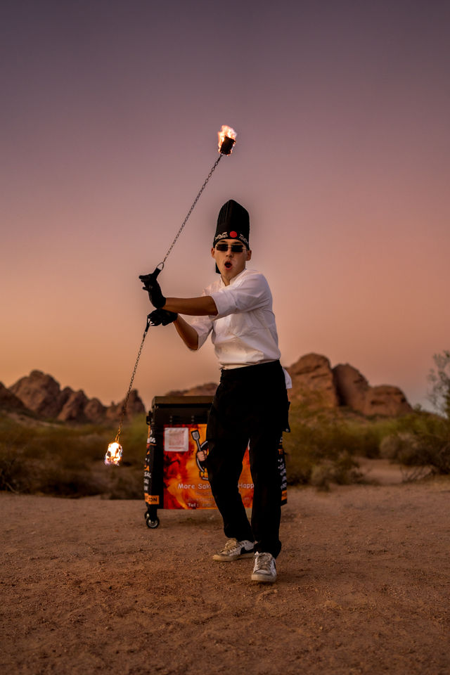 Fire-spinning chef performer in white jacket and tall hat twirls a flaming chain in a desert at sunset, wearing gloves and sunglasses with rocky hills and a food cart in the background.