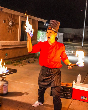 Wide-eyed chef in a red jacket and tall hat performing a flaming teppanyaki show outdoors at night, holding a ball of fire in one hand and a torch over a propane grill on a concrete patio.