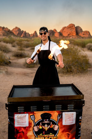 Smiling chef in apron and headband twirling flaming torches over a flat-top griddle in a desert with red rock formations at sunset