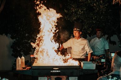 Teppanyaki chef at an outdoor hibachi grill creating a towering flame during a lively nighttime cooking performance, with diners and a camera watching nearby.