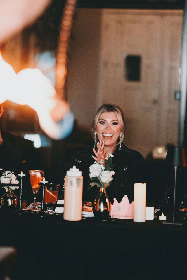 Smiling woman applauding at a candlelit restaurant dinner table with cocktails, a floral centerpiece and glowing candles — lively evening celebration