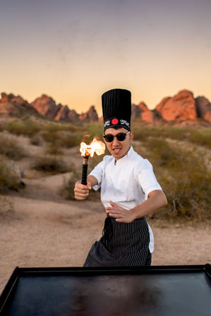 Chef in a tall black hat and striped apron performing with a flaming torch over a griddle at sunset in a red‑rock desert landscape