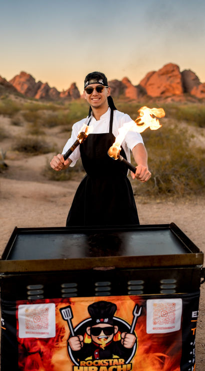 Smiling chef in apron and headband twirling flaming torches over a portable hibachi grill at a desert sunset with red rock formations in the background, outdoor hibachi fire show.