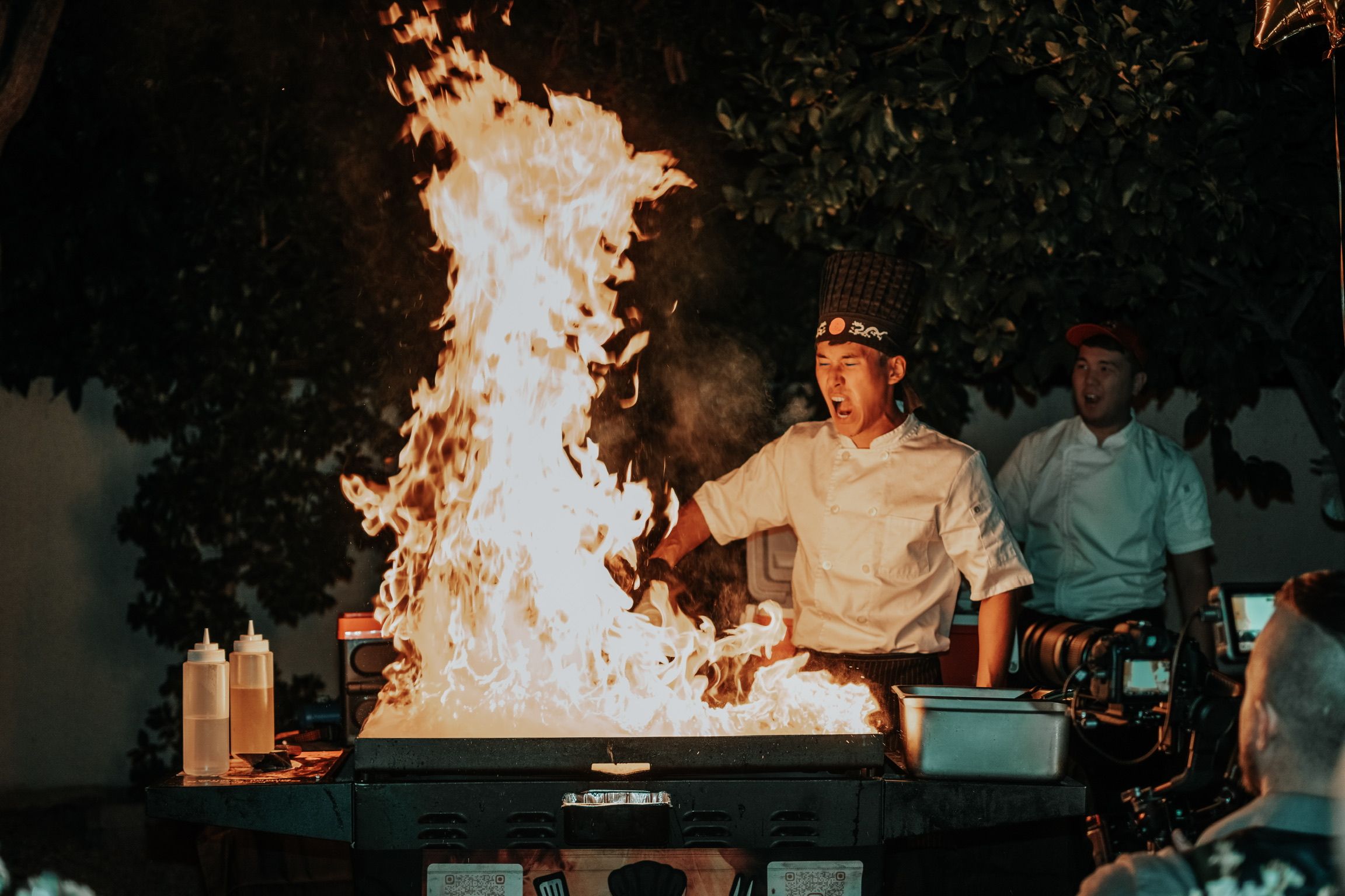 Hibachi chef in white uniform and tall hat creates towering flames on an outdoor teppanyaki grill at night, thrilling onlookers and cameras.