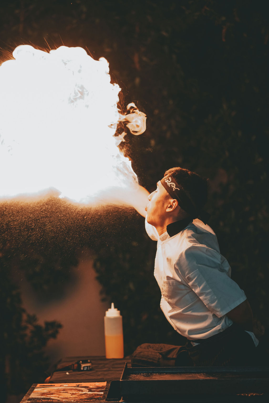 Chef performing dramatic fire-breathing at an outdoor night street-food stall, a huge plume of flame lighting up the scene