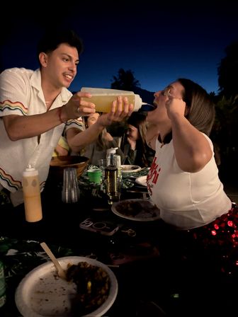 Playful backyard dinner party at night: man squirting sauce from a squeeze bottle into a friend’s open mouth while others laugh around a table with plates, cups, and palm silhouettes against the evening sky.
