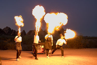 Four chefs in aprons and hats perform a night fire‑breathing show, each blowing towering flames into the twilight over a rocky desert landscape.